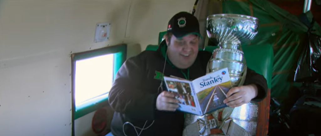 Un hombre vestido de verde sentado en un avión. En el asiento de al lado, la Stanley Cup, con el cinturón puesto. El hombre le lee una revista y la rodea con el brazo.
